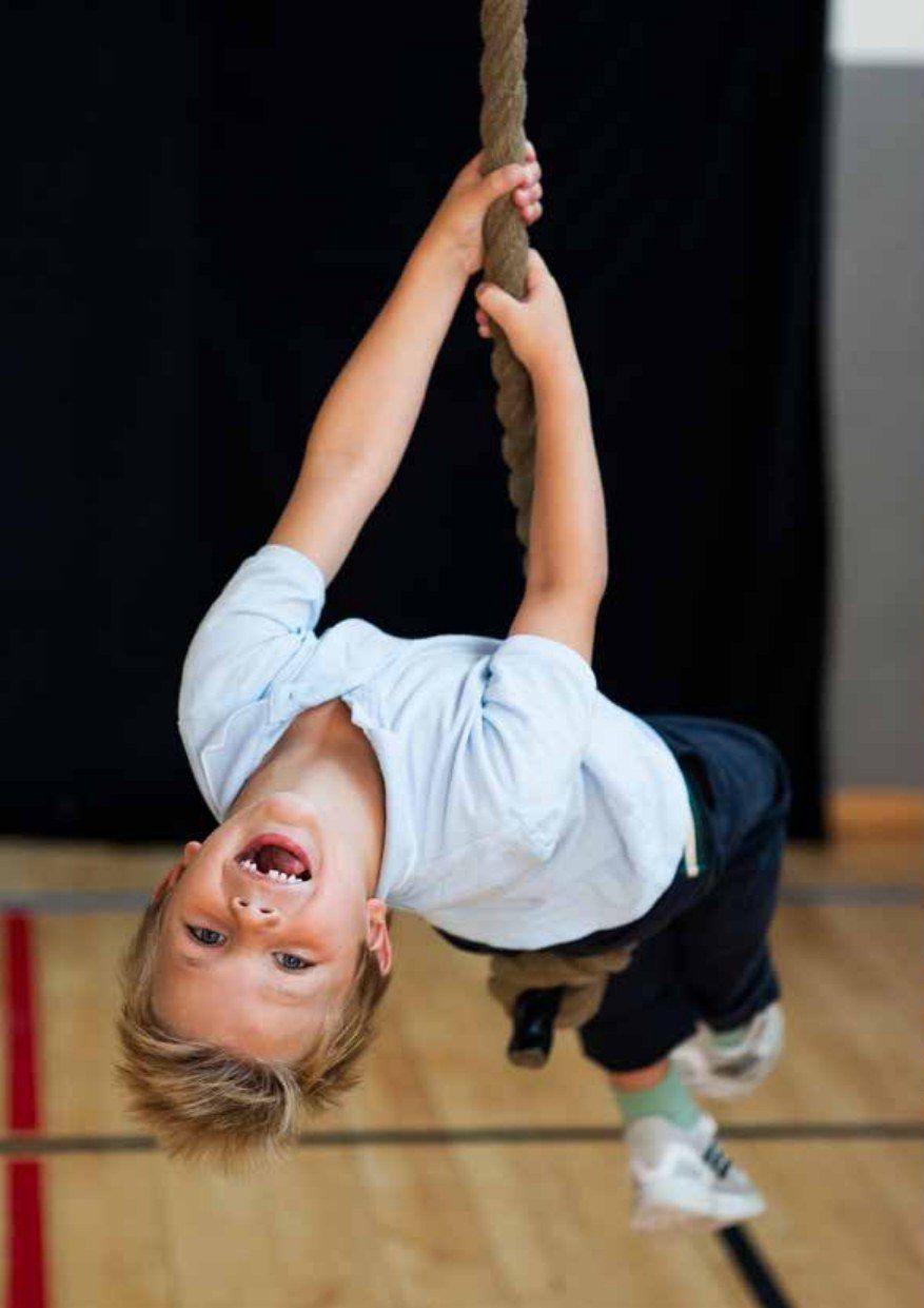 Gutt som henger opp ned i et klatretau i gymsalen og ler.
